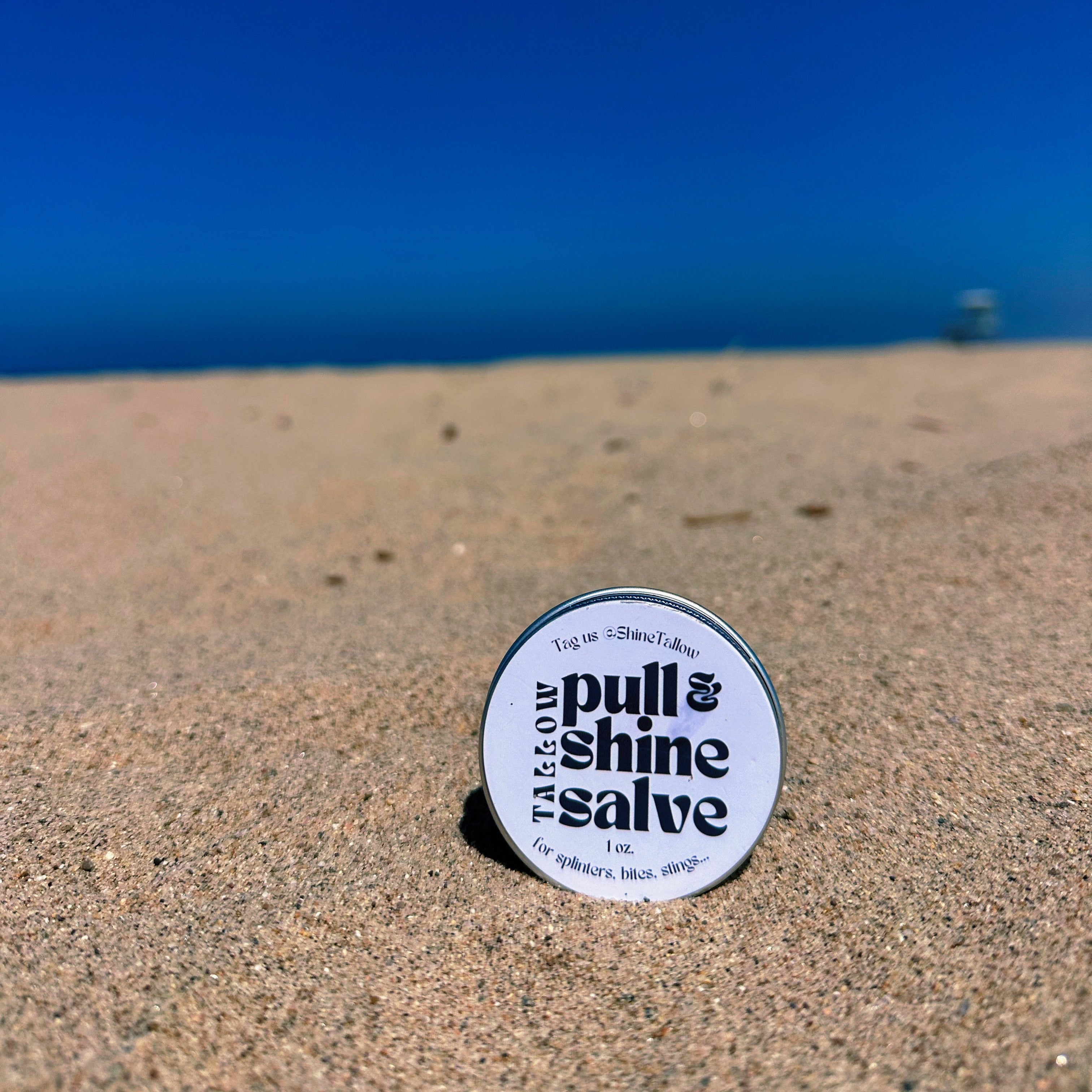 A small round tin of  tallow pull & shine salve sits on sandy beach ground with the ocean and a clear blue sky in the background.