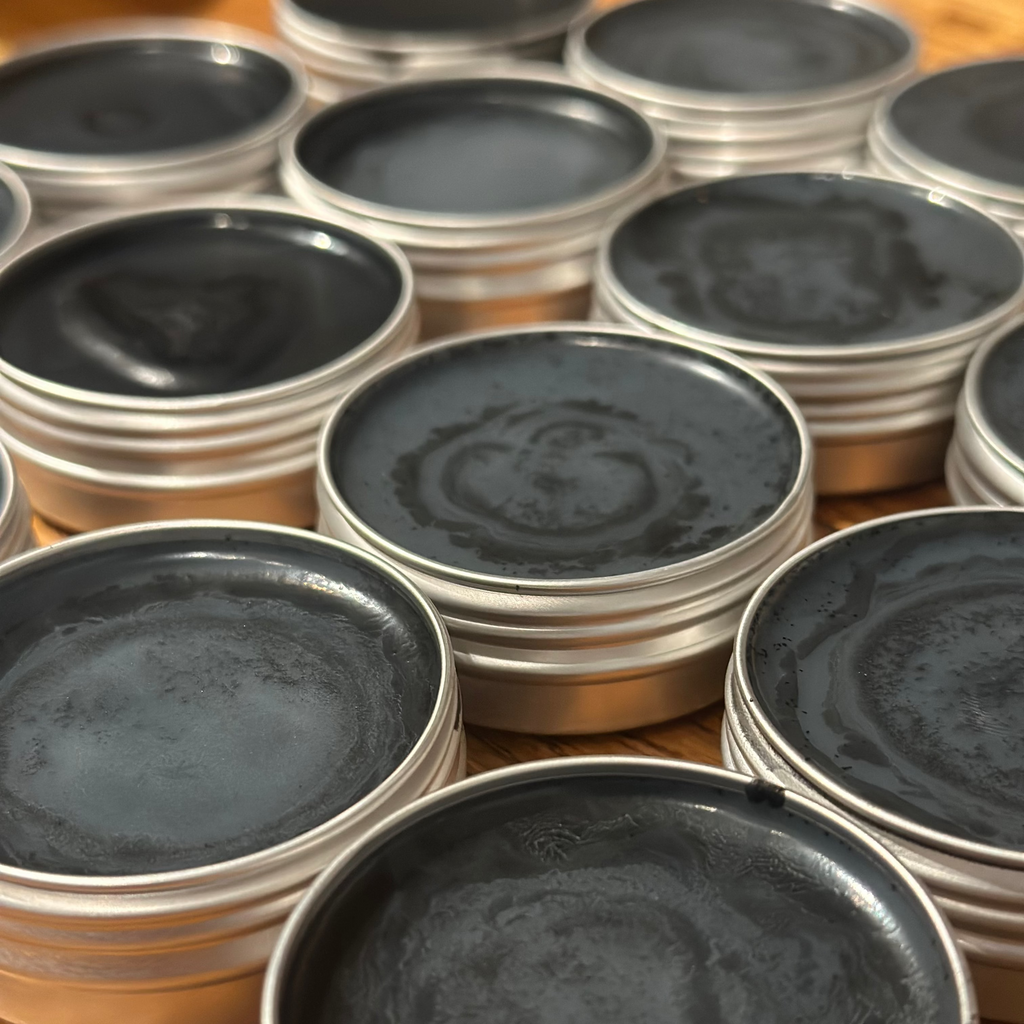Multiple round metal tins filled with charcoal salve arranged closely together on a wooden surface.