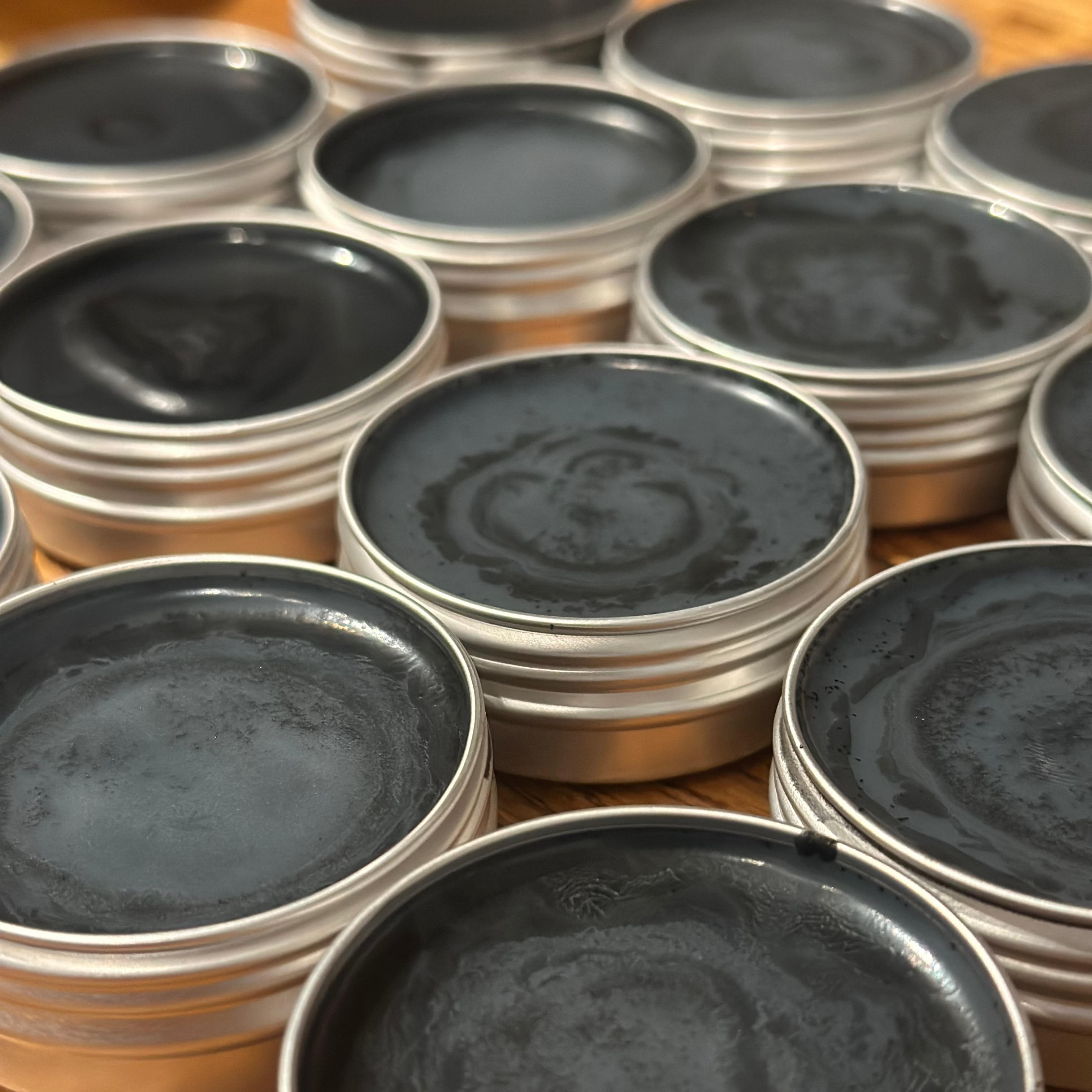 Multiple round metal tins filled with charcoal salve arranged closely together on a wooden surface.