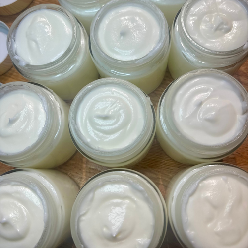 Multiple small clear jars filled with a thick tallow balm, arranged on a wooden surface. One jar is open and its lid is partly visible at the upper left