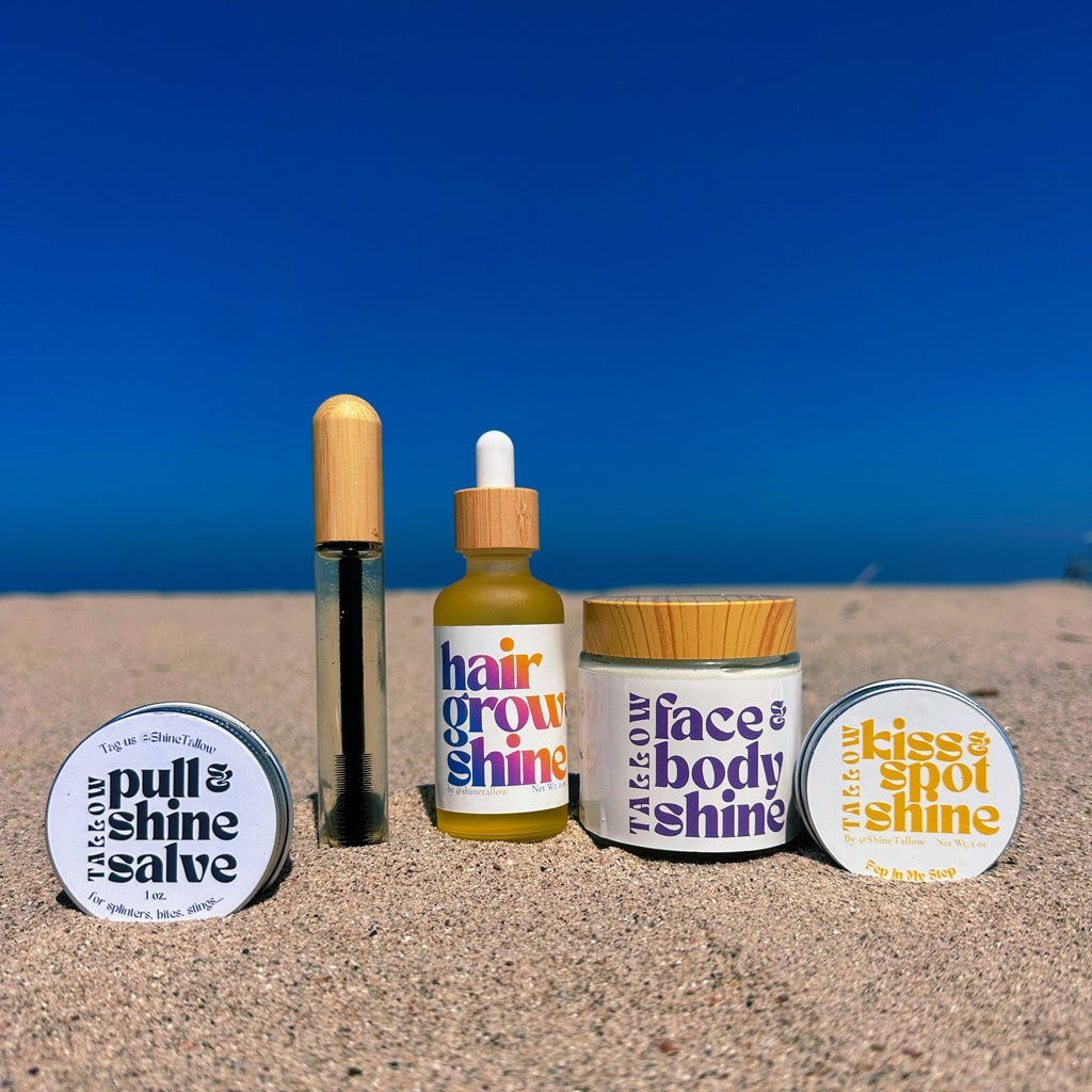 Four Tallow Beauty products arranged on sand with the ocean and blue sky behind them: a round pull & shine salve tin, a hair grow & shine dropper bottle, a face & body shine jar, and a kiss & shine salve tin.