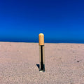 A small cylindrical product with a light wood-toned cap stands upright in sand, with a clear body showing dark liquid inside, set against a bright blue sky and ocean background.