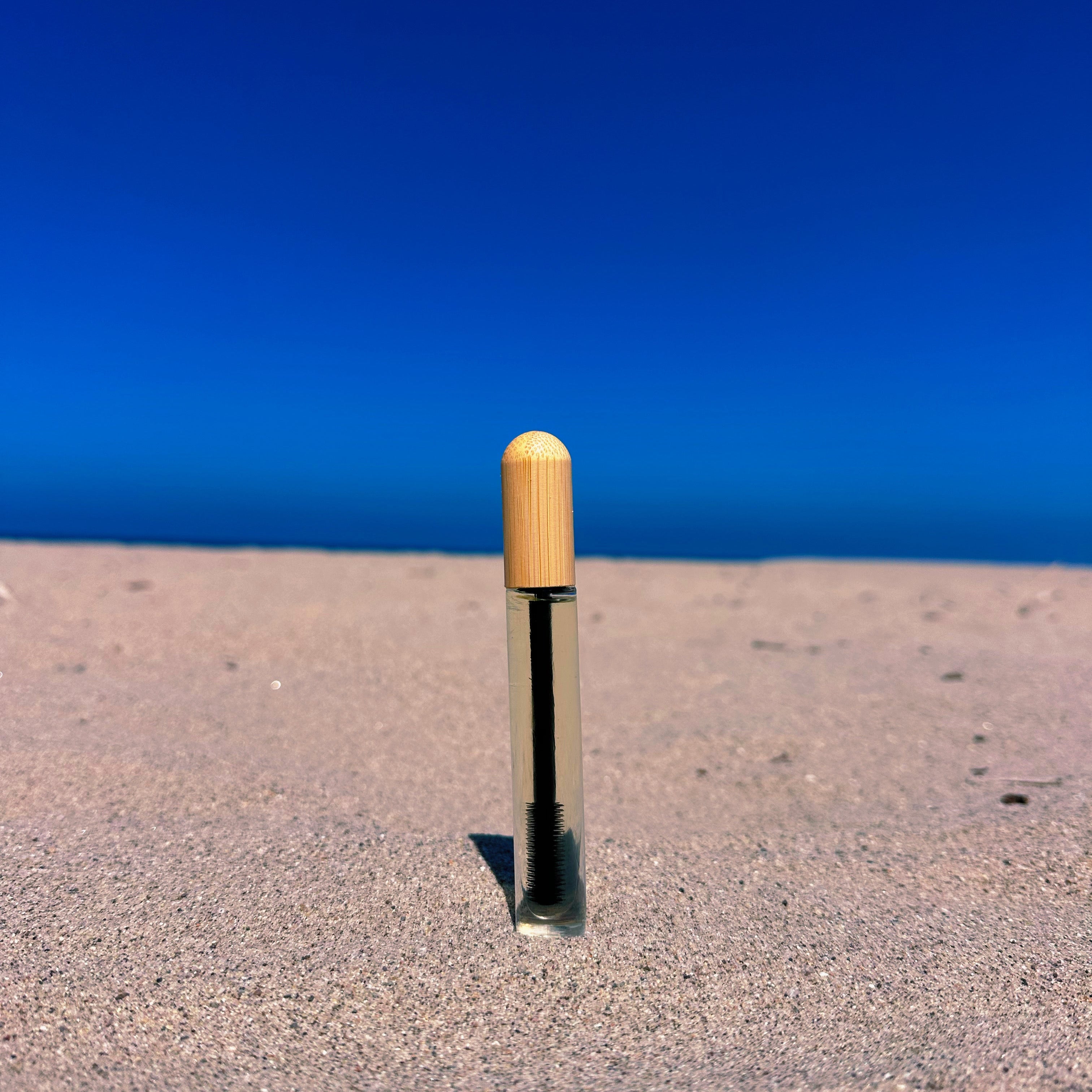 A small cylindrical product with a light wood-toned cap stands upright in sand, with a clear body showing dark liquid inside, set against a bright blue sky and ocean background.
