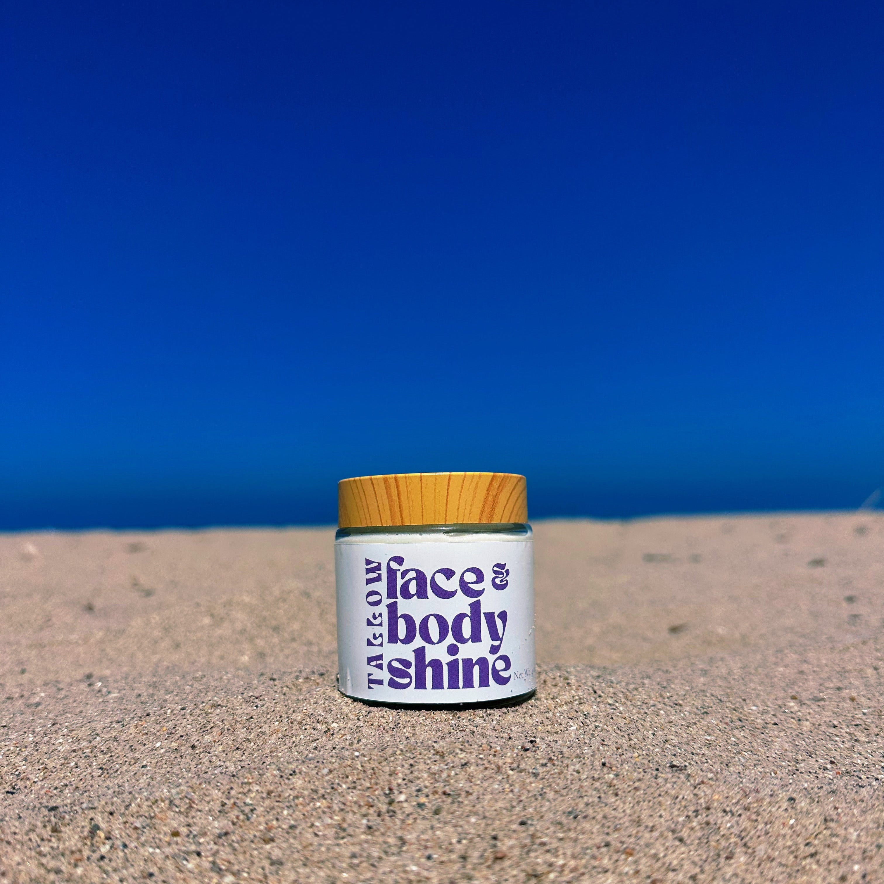 A small white jar labeled 'face & body shine' with a yellow lid sits on sandy ground with a deep blue sky background.