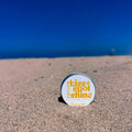 A round tin of tallow 'Kiss & Spot Shine' skin product standing upright on a sandy beach with the ocean and blue sky blurred in the background.