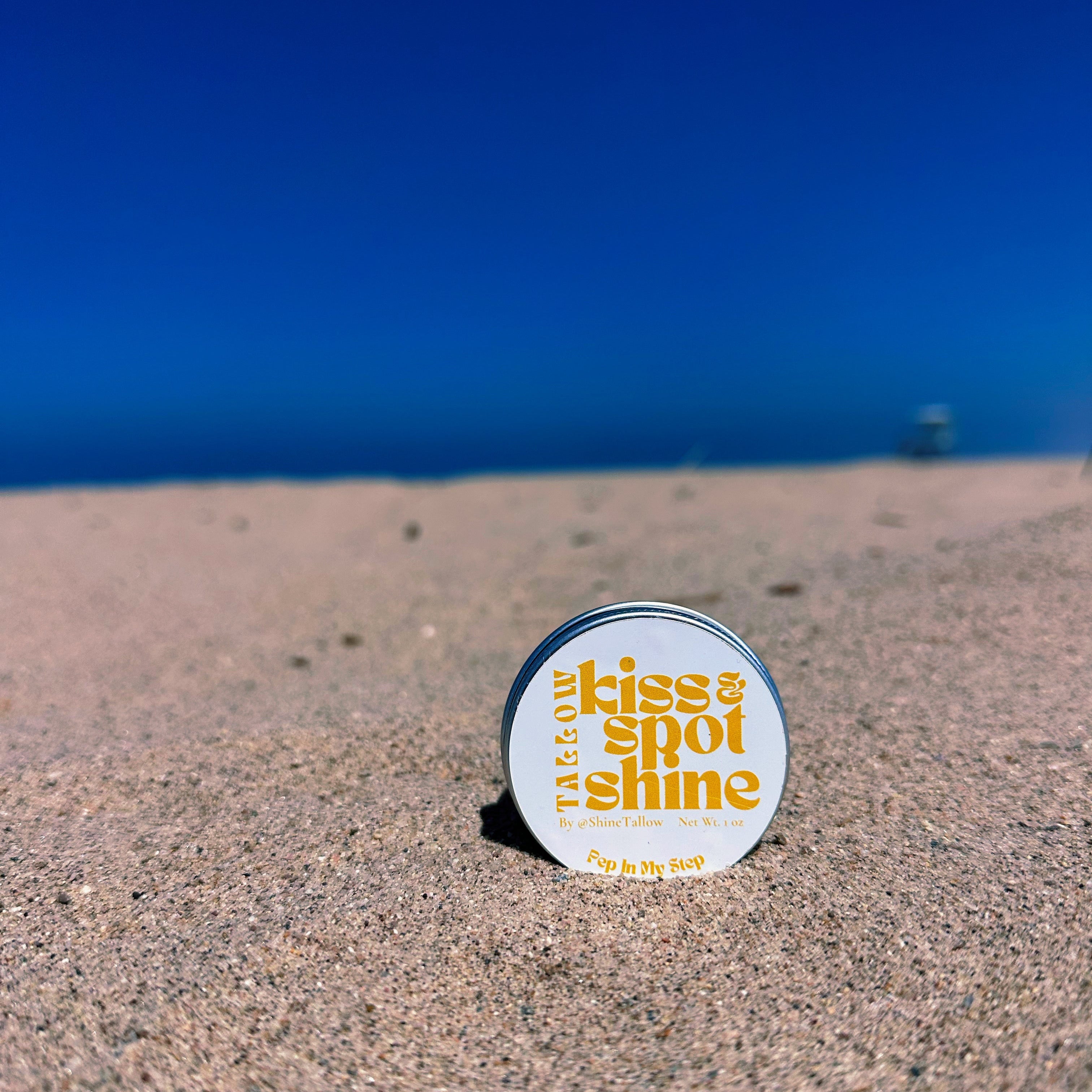 A round tin of tallow 'Kiss & Spot Shine' skin product standing upright on a sandy beach with the ocean and blue sky blurred in the background.