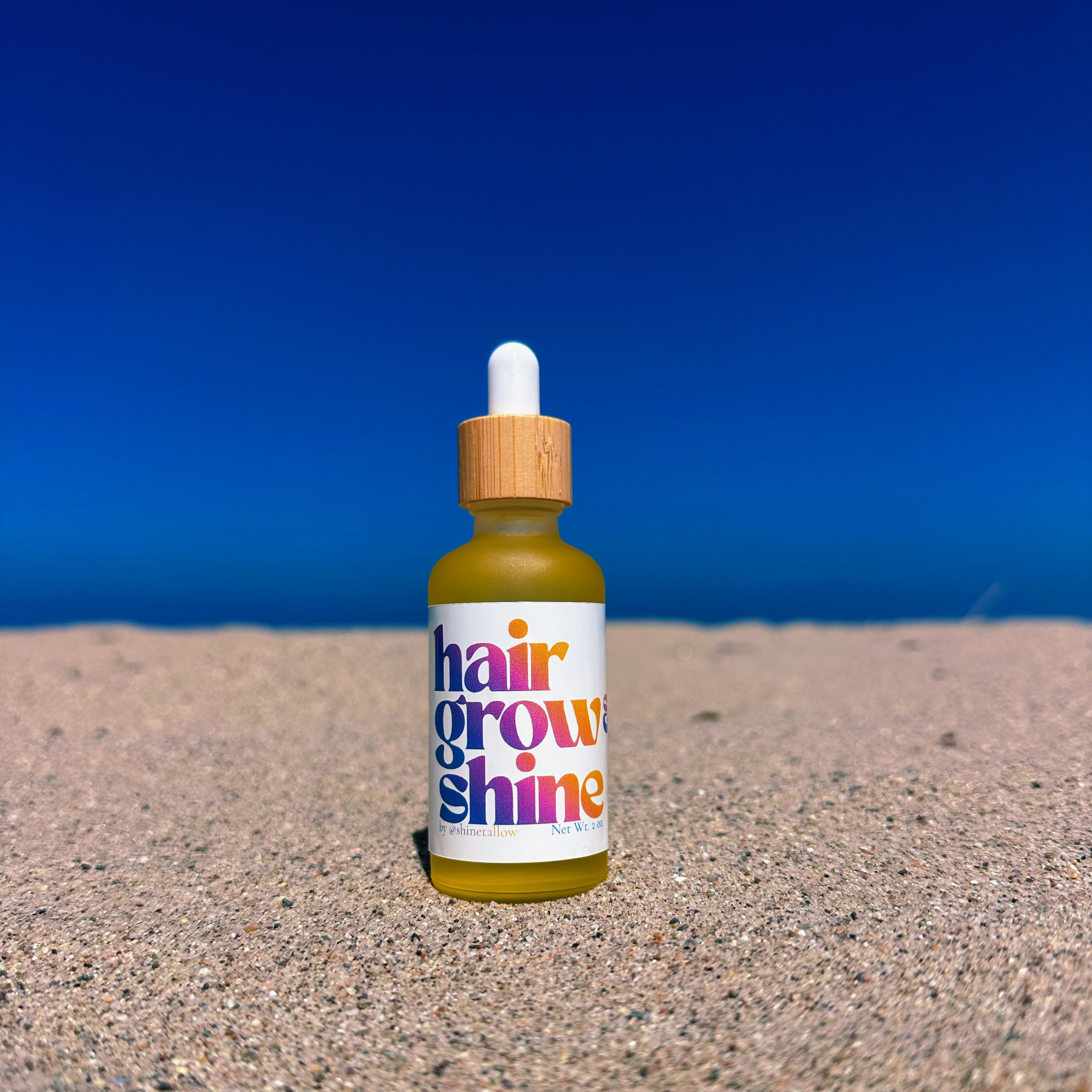 A small dropper bottle labeled 'hair grow & shine' in colorful text, standing upright on sand with a clear blue sky background.
