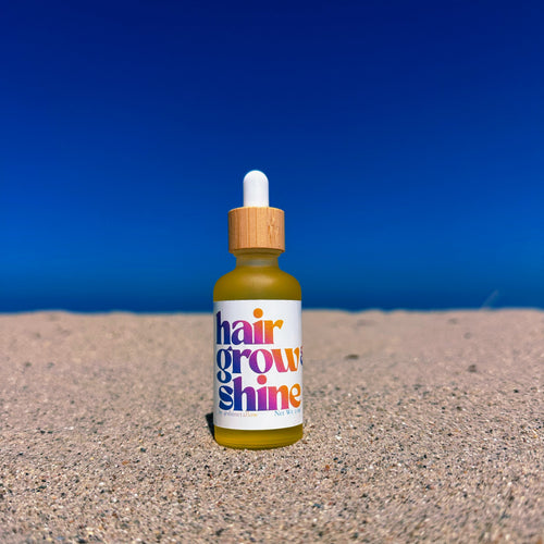 A small dropper bottle labeled 'hair grow & shine' in colorful text, standing upright on sand with a clear blue sky background.
