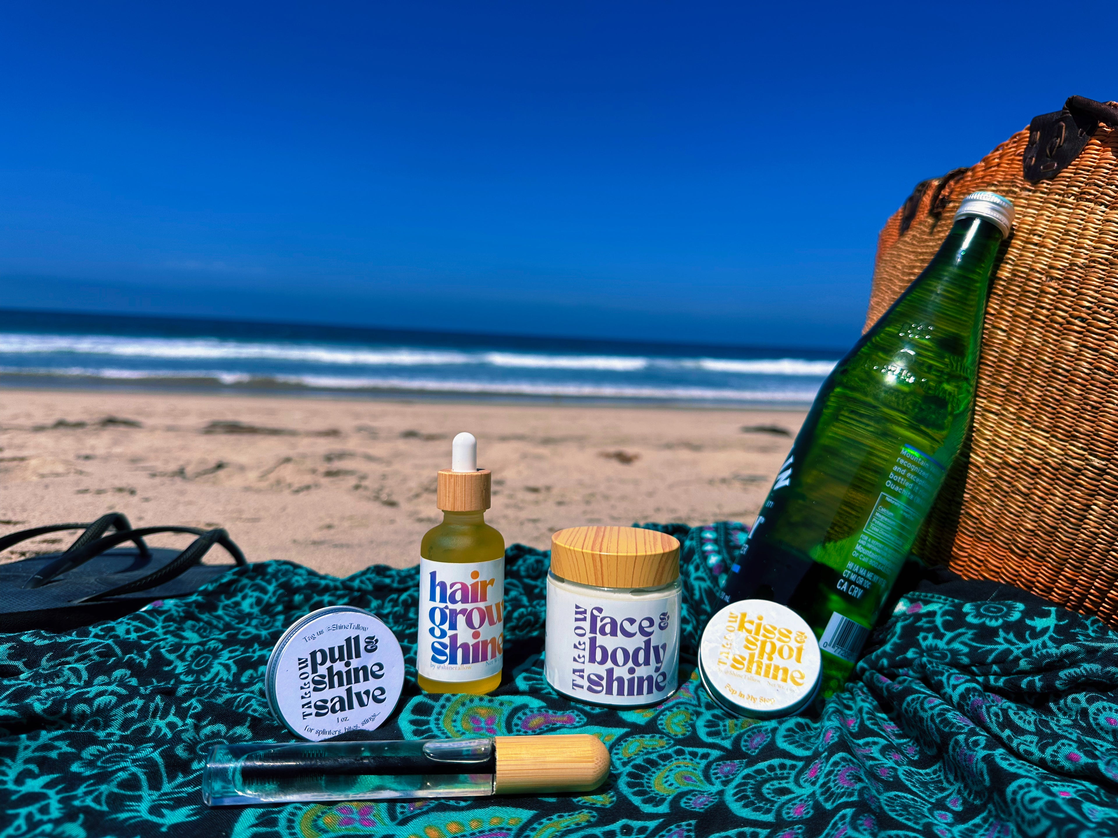 A beach-themed product arrangement on a teal patterned blanket, featuring a green glass bottle, a hair growth shine dropper bottle, a face/body shine jar, a pull & shine salve tin, and a glass vial, with a woven basket and ocean backdrop.